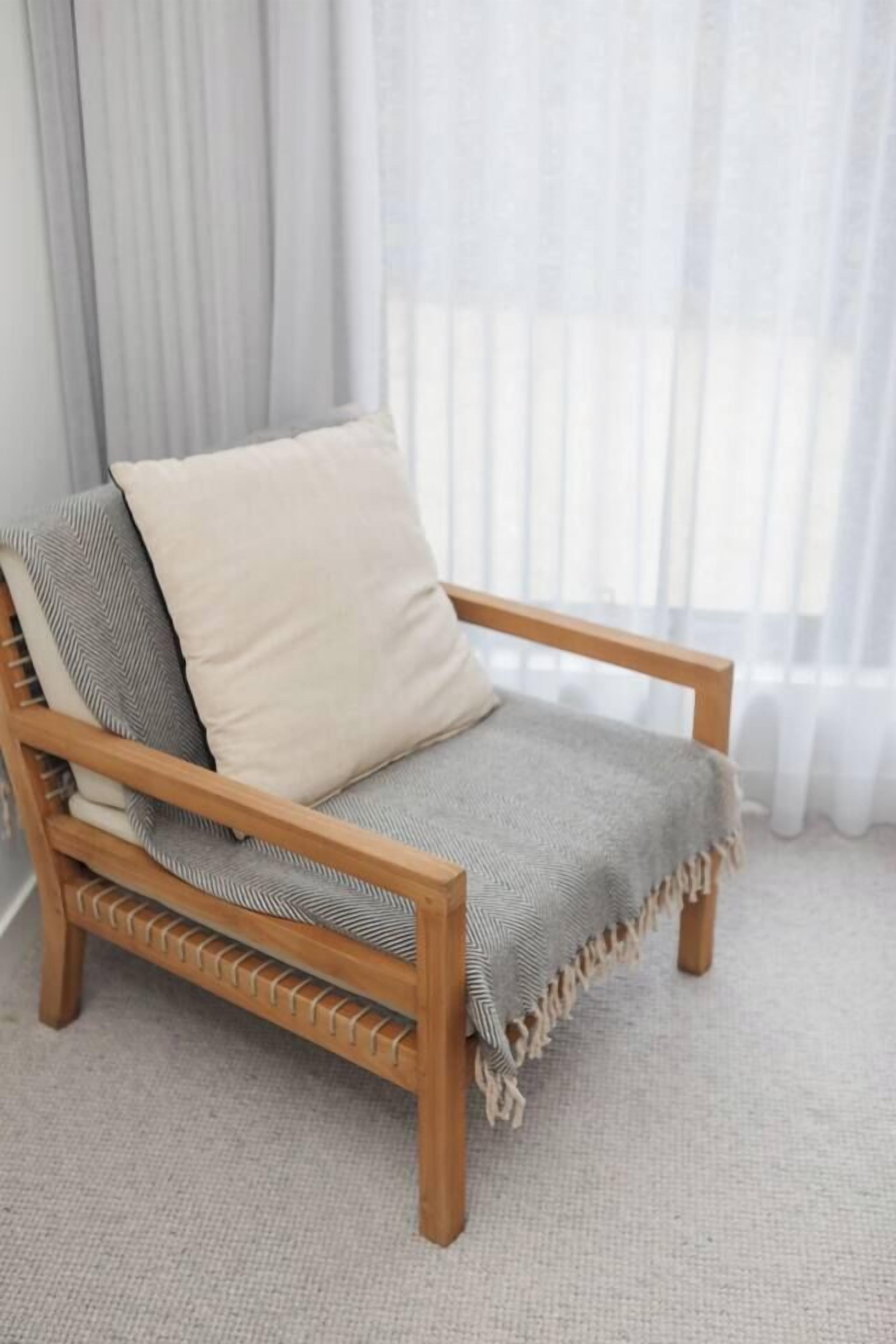 A wooden chair with a cushion and a blanket, positioned in front of a window with sheer curtains.