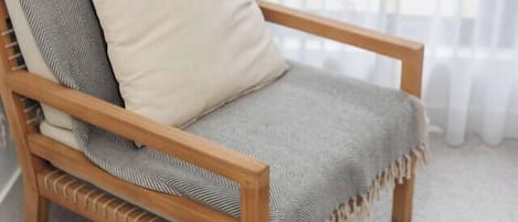 A wooden chair with a cushion and a blanket, positioned in front of a window with sheer curtains.