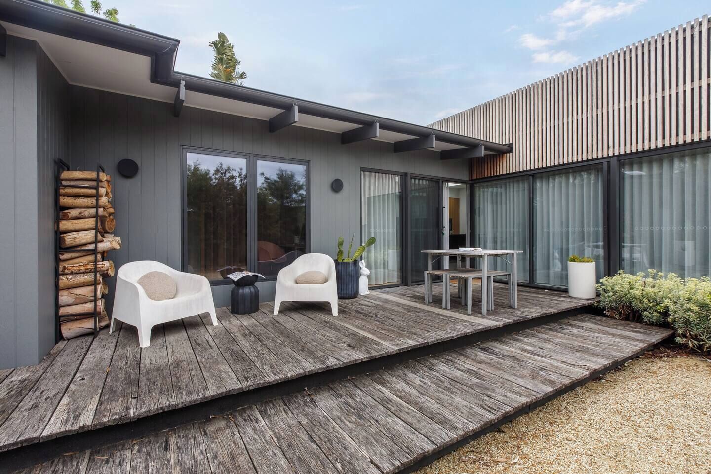A modern outdoor deck with two white chairs, a small table, and a log display.