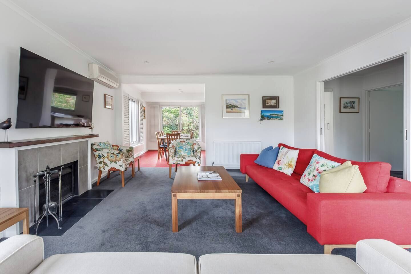 A living room with a red sofa, a wooden coffee table, and a fireplace. There is a television mounted on the wall and a dining area with chairs and a table.