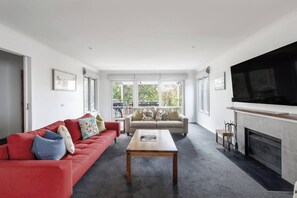 A spacious living room with a red sofa, beige sofa, and a wooden coffee table. There is a fireplace and a large TV mounted on the wall. - Melrose Nest Peaceful Mount Martha Stay near Beach (Mount Martha)