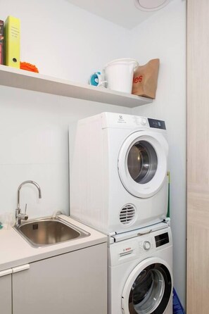 A compact laundry area with a stacked washer and dryer, a stainless steel sink, and a shelf with various items. - Bayside Calm Walk to Mornington Foreshore and Dining (Mornington)