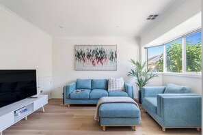 Ein modernes Wohnzimmer mit Fernseher, einem blauen Sofagarnitur, einem Couchtisch und einem großen Fenster mit Blick ins Freie.
