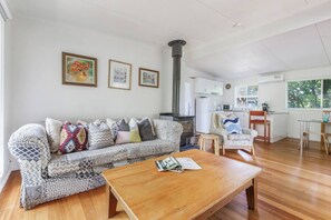 A living room with a sofa, a coffee table, and a fireplace. There are framed pictures on the wall and a kitchen area in the background. - Bayside Cottage Leafy Stay near Rye Beach and Dining (Rye)