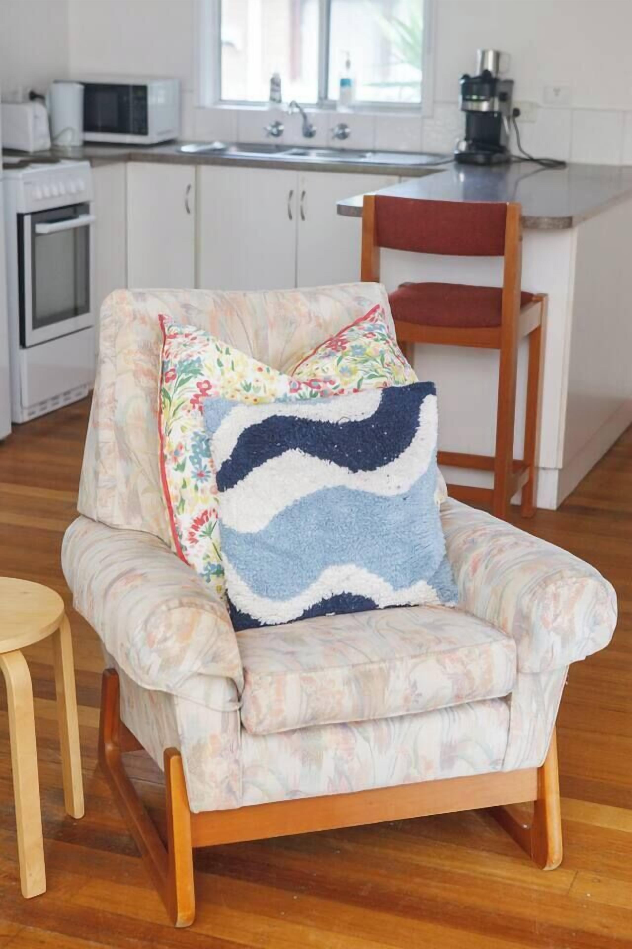 Une chaise recouverte d’un tissu à motifs floraux, avec un coussin bleu et blanc, dans une pièce dont le sol est en bois et les meubles de rangement sont blancs.