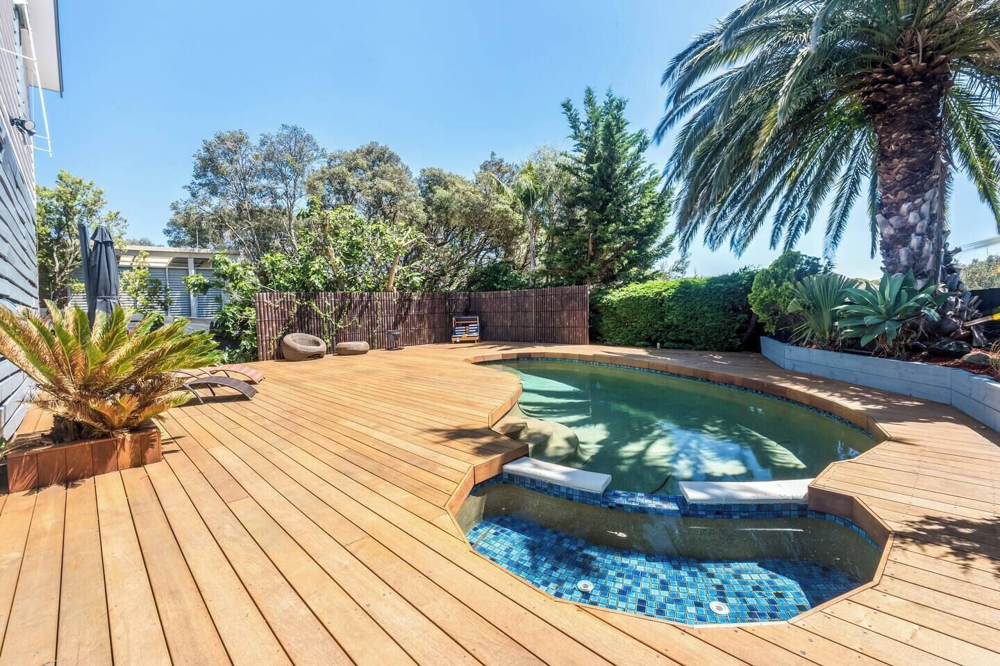 Eine Holzterrasse mit Schwimmbad, umgeben von verschiedenen Pflanzen und Bäumen.
