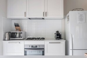 A modern kitchen with white cabinets, a microwave, a coffee maker, a kettle, and a refrigerator. - The Shack Your Ultimate Family Escape by the Sea (Rye)
