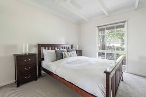A bedroom with a wooden bed, white bedding, a nightstand with drawers, and a window with a view of trees. - The Shack Your Ultimate Family Escape by the Sea (Rye)