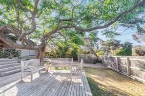 A wooden deck with benches, surrounded by a fenced yard with trees and a house in the background. - The Shack Your Ultimate Family Escape by the Sea (Rye)