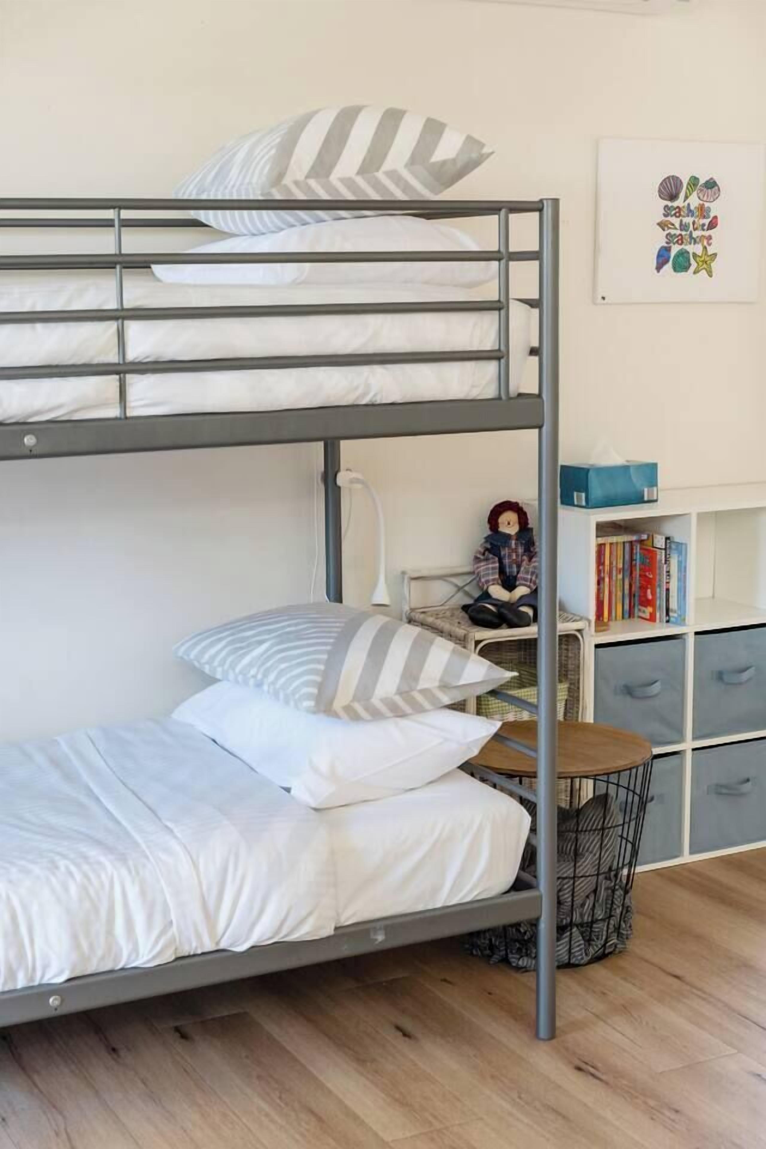 Ein Etagenbett mit weißer Bettwäsche und gestreiften Kissen, ein kleiner Schreibtisch mit Stuhl, ein Bücherregal und eine Wandlampe.