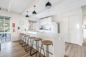 A modern kitchen with a central island, bar stools, pendant lighting, and a view of the outdoors. - The Shack Your Ultimate Family Escape by the Sea (Rye)