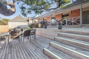 Una terraza de madera con mobiliario exterior, una escalera que lleva a una zona cubierta y un árbol grande sobre ella.