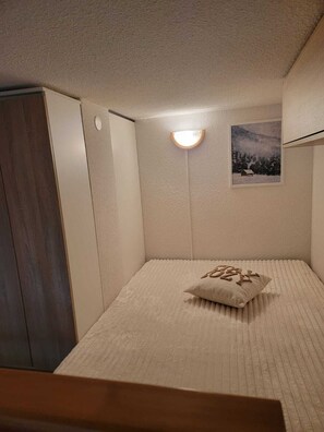 Um quarto pequeno e acolhedor com uma cama, uma mesa de cabeceira, uma luminária na parede e um quadro emoldurado.