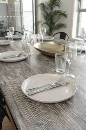 A dining table set with plates, cutlery, and glasses, with a plant and window in the background. - Seventh and Shore Spacious Poolside Group Getaway (Rosebud)