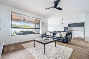 A modern living room with a large window, a black leather sofa, a coffee table, and a kitchen area in the background. - Seventh and Shore Spacious Poolside Group Getaway (Rosebud)