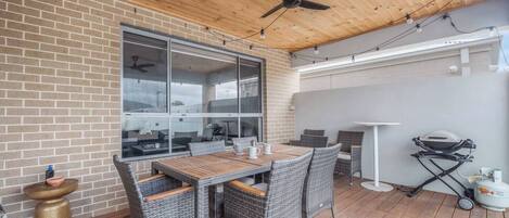 Un patio exterior con pared de ladrillo, ventilador de techo, mesa con sillas y una parrilla.