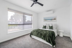 Ein Schlafzimmer mit großem Fenster, Deckenventilator, Bett mit grüner Bettdecke, Nachttisch und Wand-Klimaanlage.