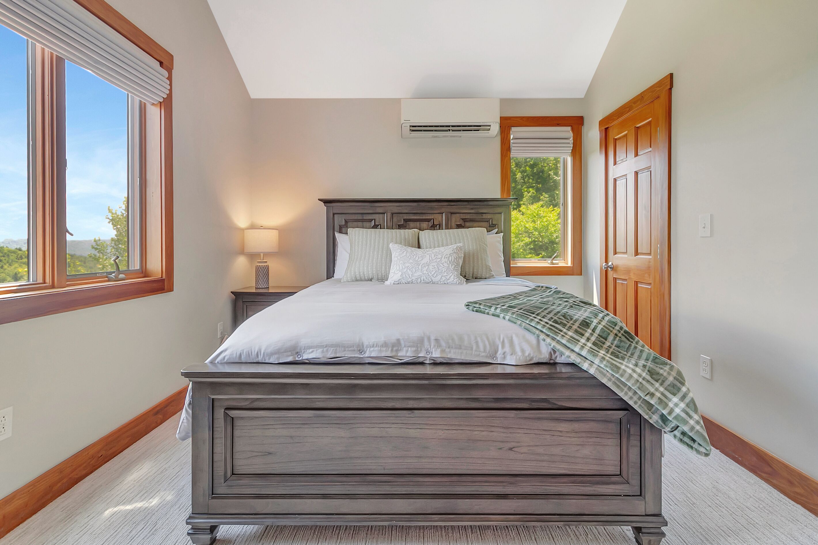 A bedroom with a large bed, a nightstand, a lamp, an air conditioning unit, and a view of greenery outside.