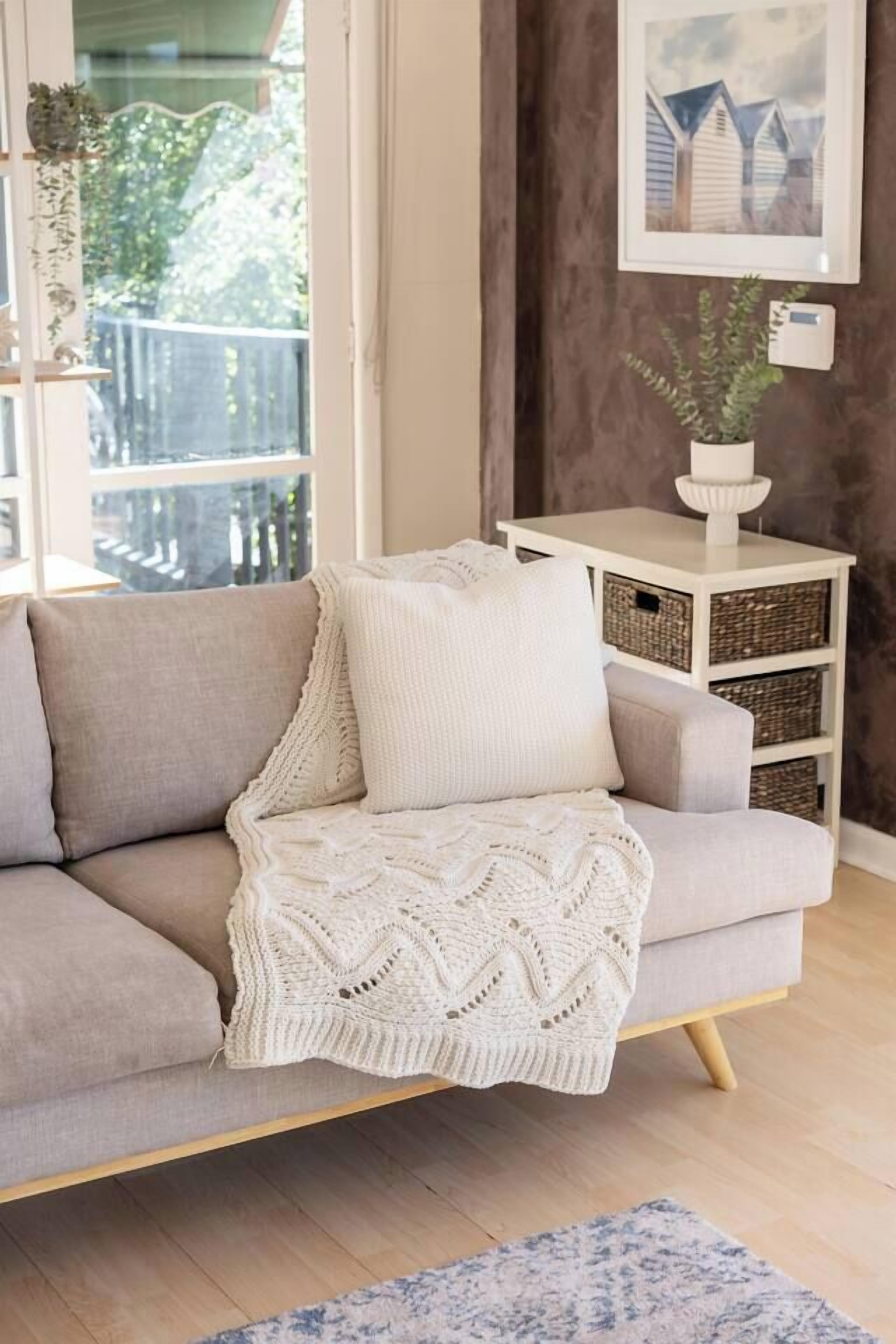 Een woonkamer met een bank, een gebreide plaid, een witte bijzettafel met manden en een lijst met een foto aan de muur.