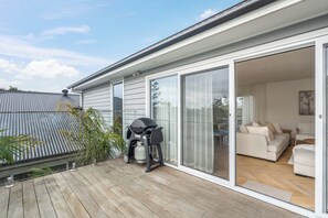 A modern house with a wooden deck, a barbecue grill, and a sliding glass door leading to a living room with a sofa and a coffee table. - Reverie Poolside Haven near Beaches and Hot Springs (Rye)