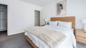 Un dormitorio con cama, mesita de noche, lámpara, un cuadro en la pared y un armario con una percha.