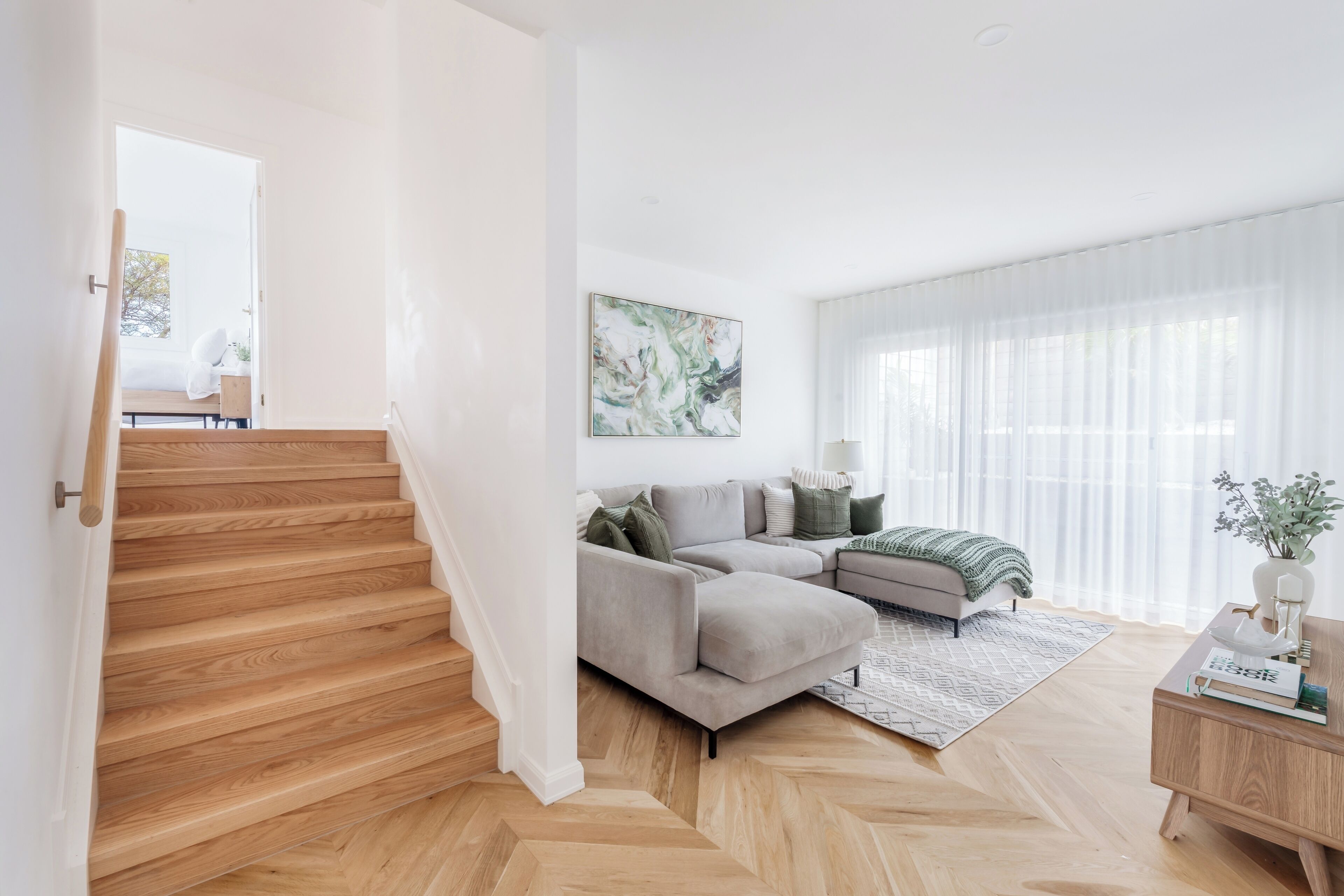 A modern living room with a herringbone wood floor, a grey sectional sofa, a large abstract painting, and a wooden coffee table.