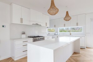 A modern kitchen with white cabinetry, a central island, a gas stove, and two hanging wicker pendant lights. - Reverie Poolside Haven near Beaches and Hot Springs (Rye)