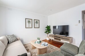 A modern living room with a sofa, a coffee table with books and a plant, a TV on a wooden stand, and two framed pictures on the wall.