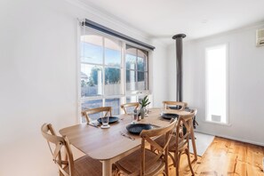 A dining area with a wooden table and chairs, large windows, and a view of the outdoors.
