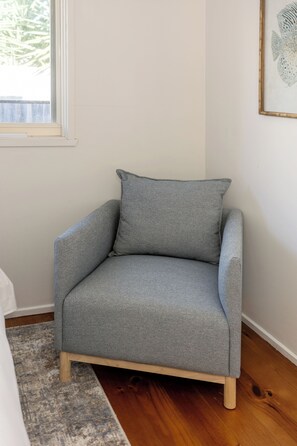 A grey armchair with wooden legs, positioned in a corner of a room with a wooden floor and a white wall.