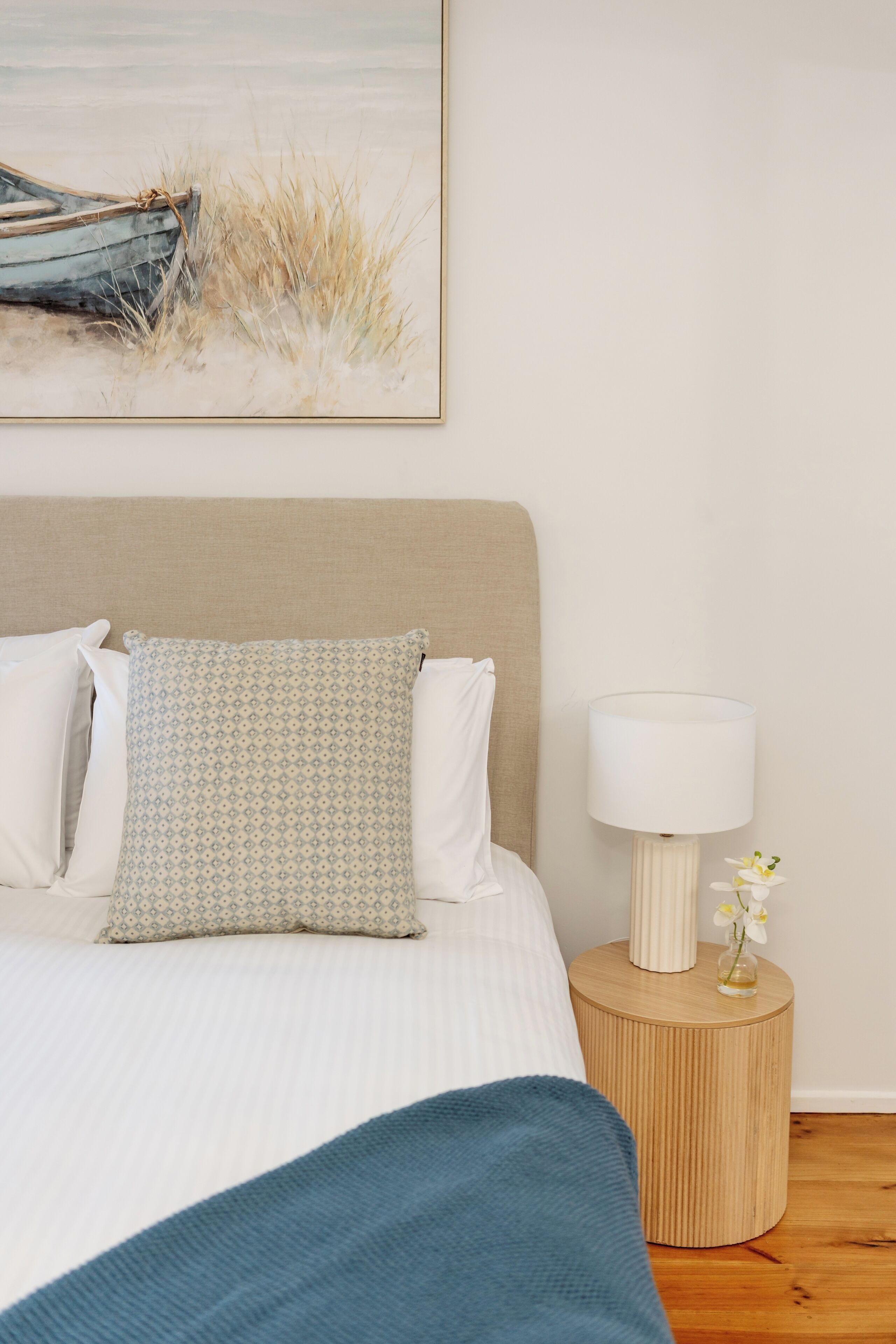 Un lit bien fait, agrémenté d’un coussin à motifs, d’une lampe de chevet et d’un petit vase avec des fleurs, posé sur une table de chevet en bois.
