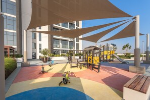 A playground with a slide and climbing structures, surrounded by a modern building with large windows. - Stayshort - Minutes to Downtown - Sleek Meydan Apartment With Balcony - Sleeps 4 (Dubai)