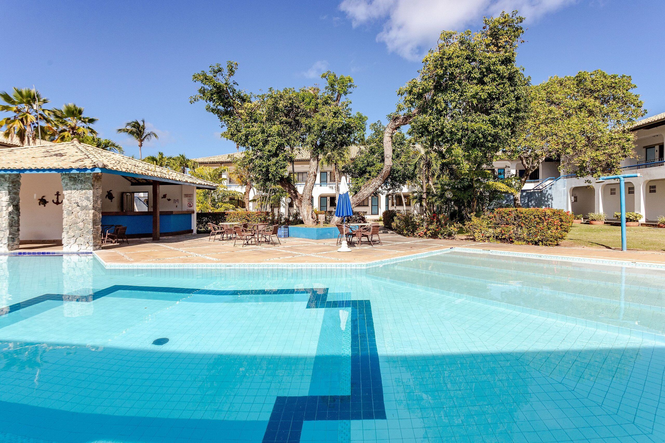 Una piscina con sillas de descanso y mesas, rodeada de edificios y árboles.