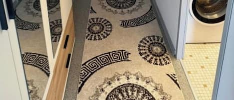 A narrow hallway with a patterned carpet leading to a door, a washing machine in the adjacent room, and a tiled floor.