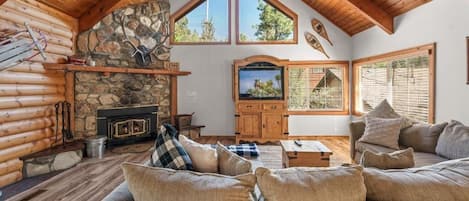 A log cabin interior with a stone fireplace, wooden ceiling beams, a television, and a cozy seating area.