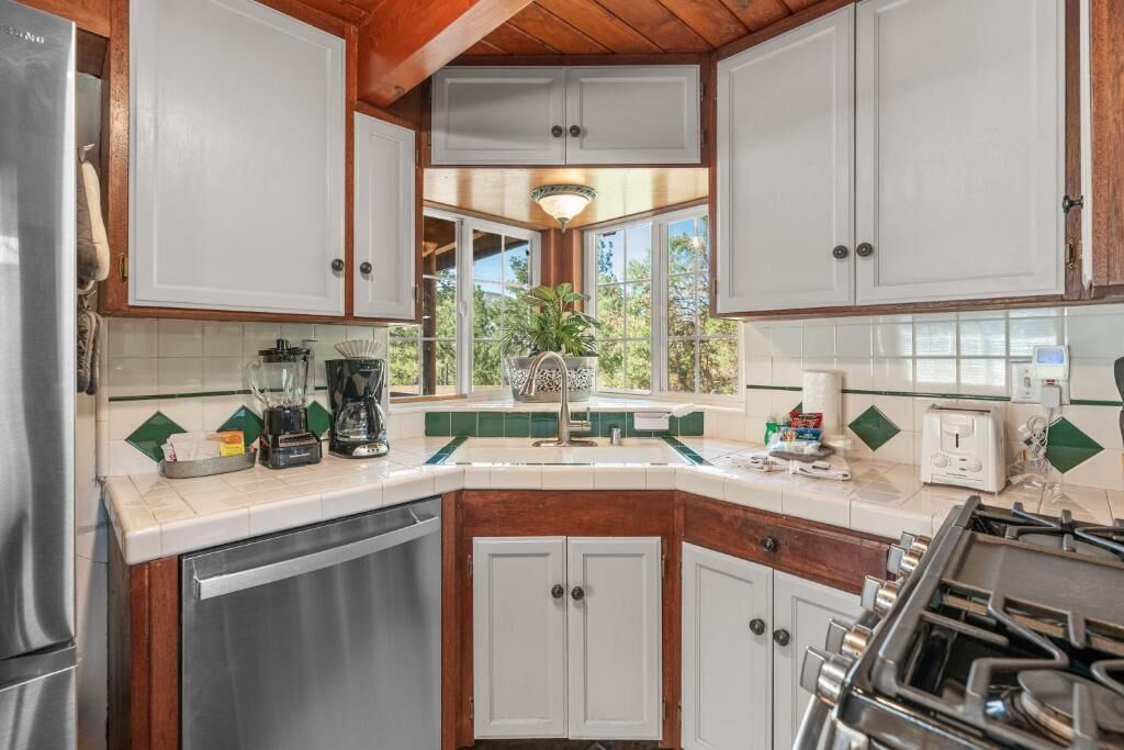 Una cocina con armarios blancos, estufa, lavavajillas y ventana con vistas al verdor.
