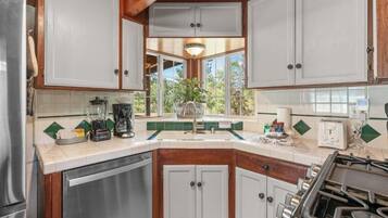 Una cocina con armarios blancos, estufa, lavavajillas y ventana con vistas al verdor.