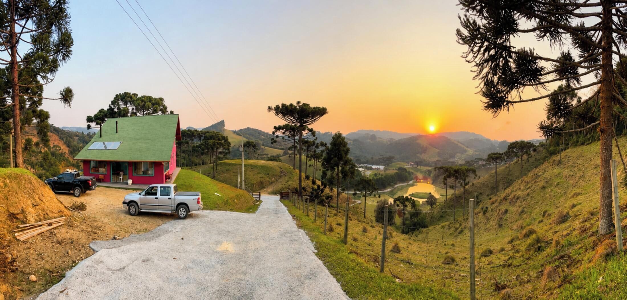 Un camino rural que lleva a una casa roja con techo verde, rodeada de colinas y un atardecer.