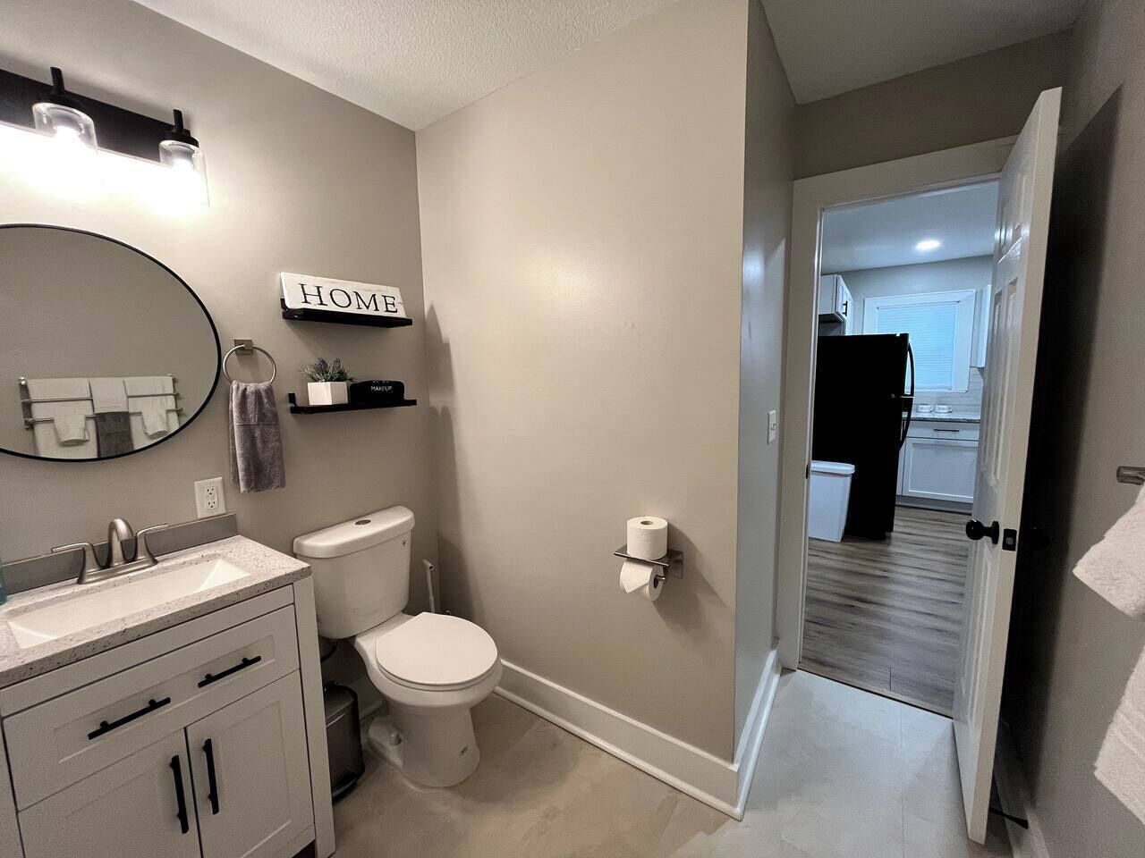 Ein Badezimmer mit Toilette, Waschbecken und Spiegel. Über dem Spiegel hängt ein Schild mit der Aufschrift 'HOME' und eine Handtuchstange ist an der Wand befestigt.