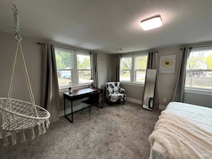 Una habitación con cama, escritorio, silla y una mecedora colgante. Hay ventanas con cortinas, una lámpara en el techo y un cuadro en la pared.