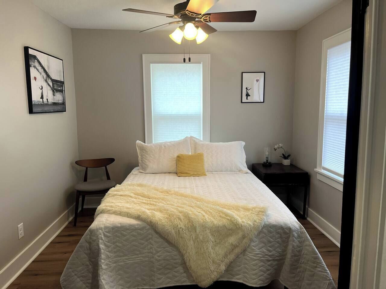 Ein Schlafzimmer mit einem Bett, Deckenventilator, Fenster, Nachttisch und einem Sessel.