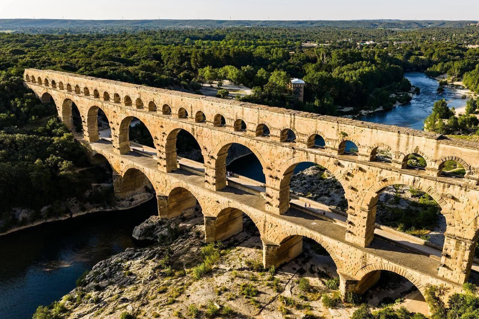 A stone aqueduct with multiple arches spanning a river, surrounded by greenery and a hilly landscape.