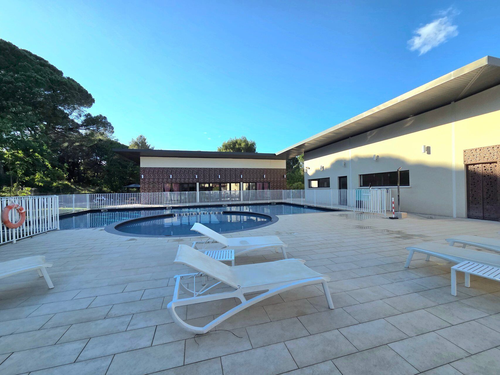 Une piscine dotée de chaises longues et entourée d’une clôture, le tout bordé par un bâtiment avec une terrasse carrelée.