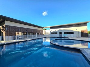 Una piscina con cerco blanco, un edificio de diseño moderno y un cielo azul despejado.