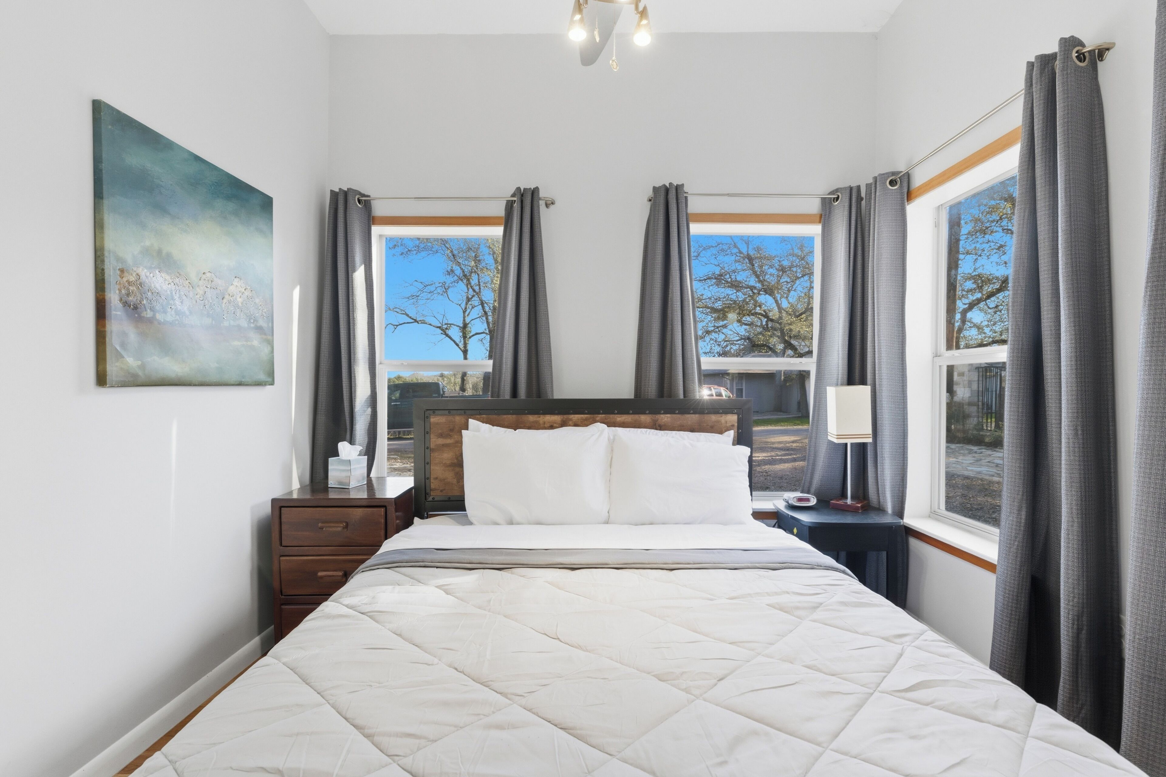 A bedroom with a bed, bedside tables, a nightstand, a painting, and a window with curtains.