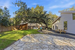 Ein Garten mit einer Steinterrasse, einem Holzzaun, einem Baum und einem Haus mit Picknicktisch.
