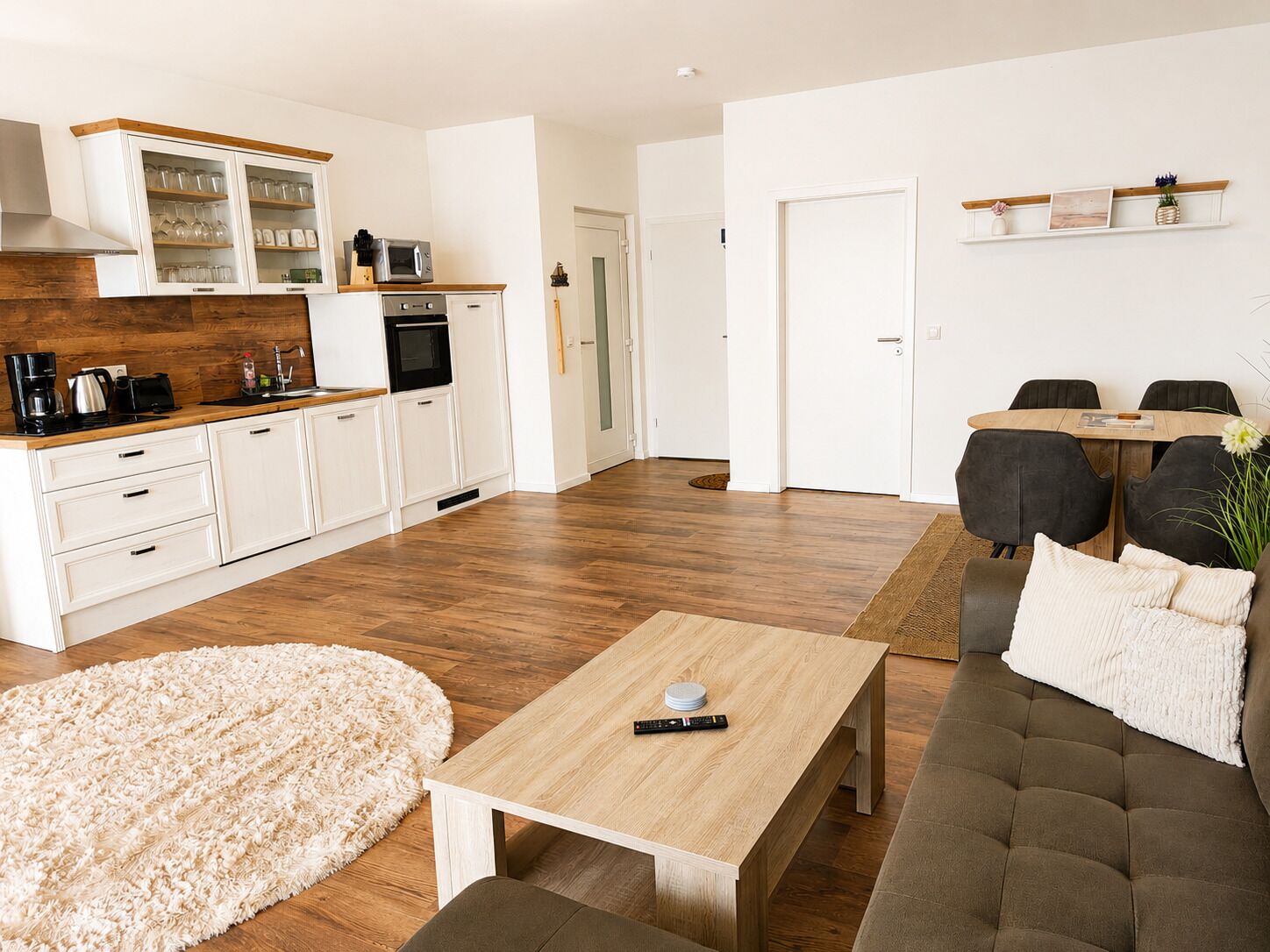 A modern kitchen with white cabinets, a microwave, and a coffee maker. There is a dining area with a wooden table and chairs.
