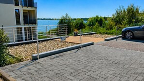 Un espace de stationnement avec une surface goudronnée, une voiture et un bâtiment doté d’un balcon donnant sur une étendue d’eau.