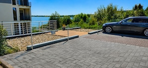 A parking area with a paved surface, a car, and a building with a balcony overlooking a body of water.