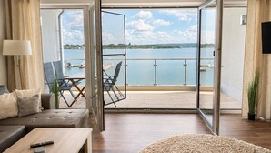 Un salon moderne avec un canapé, une table basse et des fauteuils, un balcon offrant une vue sur l’eau et les bateaux, et un téléviseur fixé au mur.